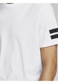 Camiseta de algodón blanca con rayas dobles negras en la manga izquierda. Textura suave, diseño de cuello redondo, mangas cortas.