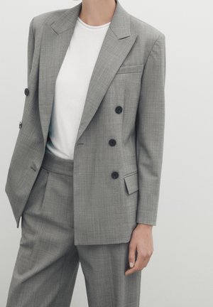 Grijze dubbelzijdige blazer van een gestructureerde stof, met grote revers en zwarte knopen, gecombineerd met een wit T-shirt en bijpassende broek.