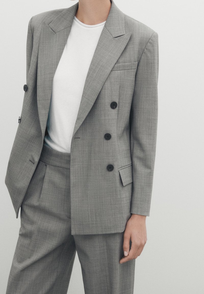 Grijze dubbelzijdige blazer van een gestructureerde stof, met grote revers en zwarte knopen, gecombineerd met een wit T-shirt en bijpassende broek.