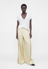 Model wearing loose cream wide-leg trousers and a white short-sleeve V-neck top, standing against a plain white background.