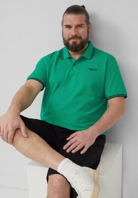 Groene polo met contrasterende marineblauwe afwerking, korte mouwen en een kraag. Draagt met zwarte shorts en witte sneakers. Eenvoudig, casual ontwerp.