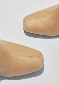 Stivaletti alla caviglia in suede color beige con punta appuntita. Cuciture visibili lungo la cucitura. Texture liscia con una leggera lucentezza.