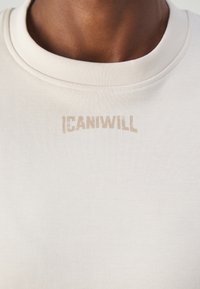 Krémové bavlněné tričko s ribbovaným kulatým výstřihem a béžovým nápisem "ICANIWILL". Hladký materiál a minimalistický design.