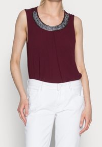 Burgunderröd ärmlös topp med en samlad detalj och en glansig, dekorerad krage. Kombineras med vita tajta jeans.