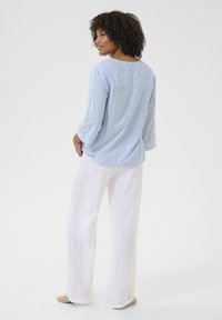 Chemise à rayures bleues et blanches avec des manches trois-quarts, associée à un pantalon en lin blanc ample, portée avec des ballerines beiges.