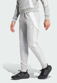 Grijze sportbroek met witte zijpanelen en drie witte strepen. Heeft een taps toelopende pasvorm en een zachte textuur. Gecombineerd met zwarte sportschoenen.
