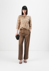 Beige bestickte Bluse mit Dreiviertelärmeln, kombiniert mit braunen Satinhosen und einer schwarzen Handtasche; das Modell trägt schwarze Riemchensandalen.