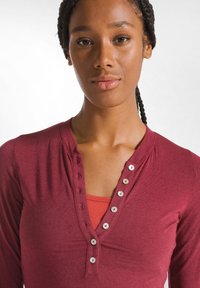 Bordeauxfarbenes langärmliges Oberteil mit einem V-Ausschnitt und silbernen Knopf-Akzenten, das über einem roten Trägershirt getragen wird.