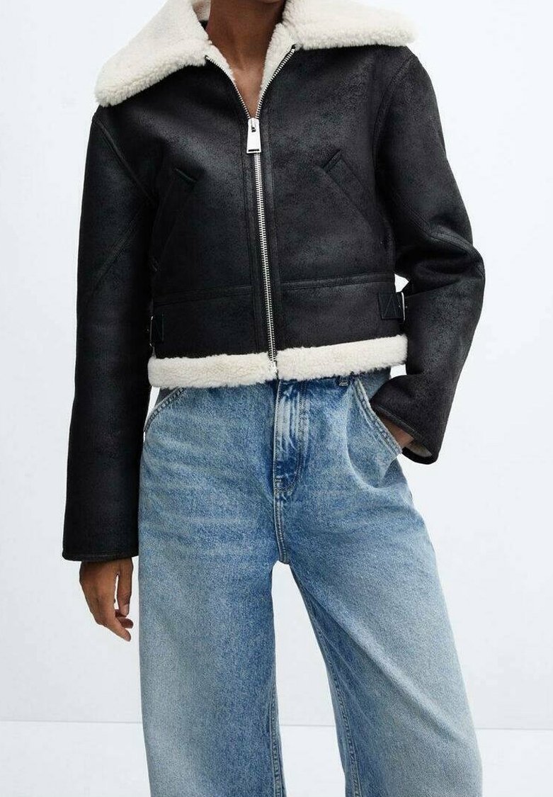 Sort cropped jakke med shearling-krave, med en frontlynlås, glat tekstur og strukturerede skuldre. Parret med lyseblå jeans.
