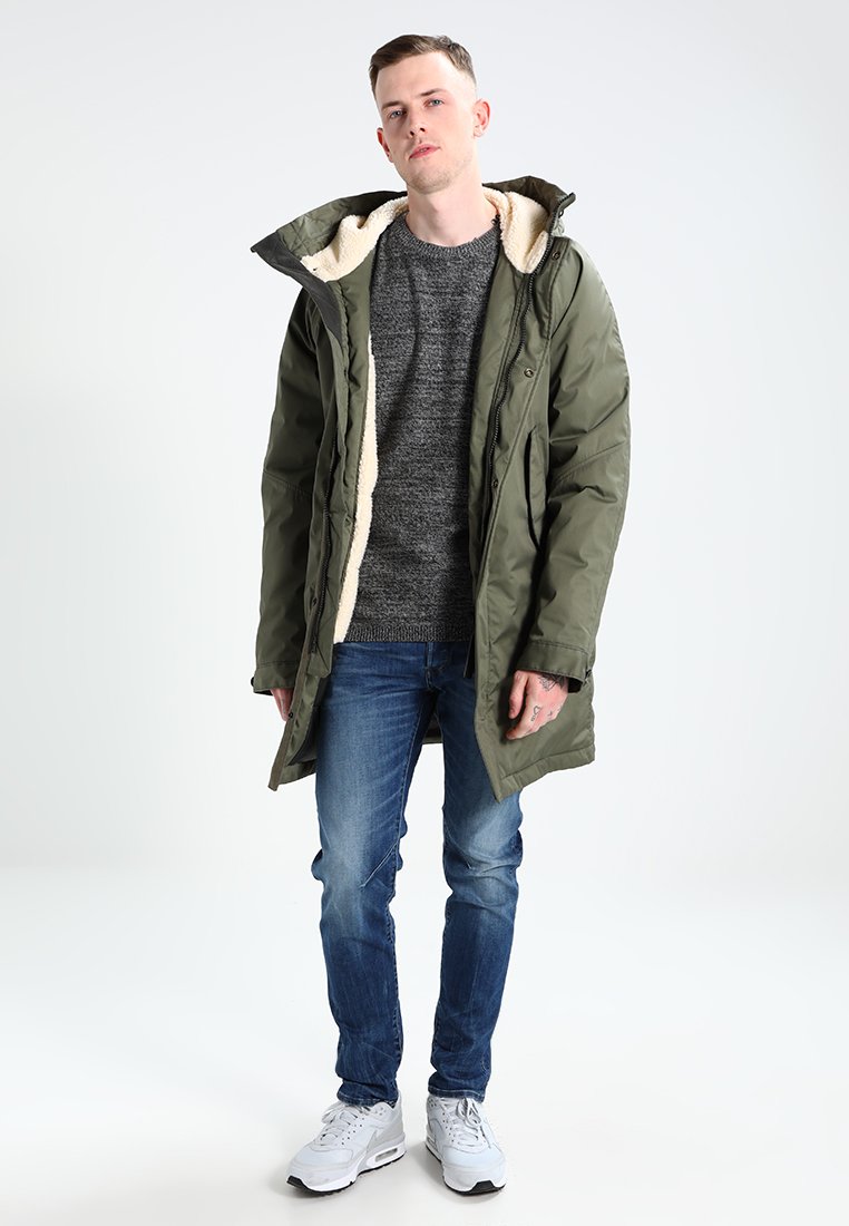 Parka matelassée vert olive avec une capuche en fourrure, assortie à un sweat gris et un jean bleu. Le modèle se tient devant un fond uni.