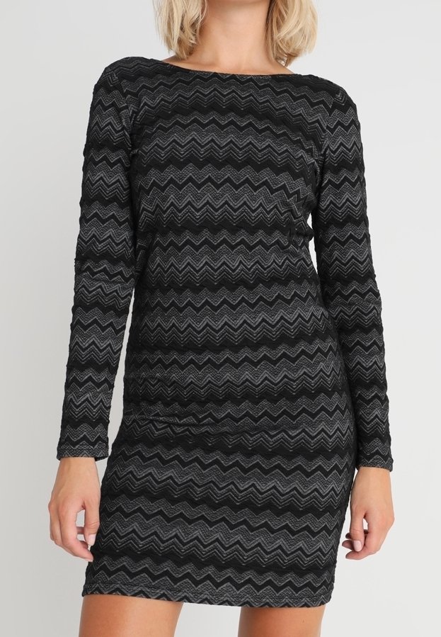 Kobieta w dopasowanej czarnej sukience z długimi rękawami, zdobionej wzorem w zygzaki w odcieniach ciemnej szarości.