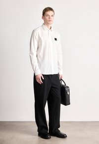 Λευκό πουκάμισο με κουμπιά και τσέπη στην πρόσοψη, συνδυασμένο με άνετο μαύρο παντελόνι και μαύρα παπούτσια, κρατώντας μια δομημένη μαύρη τσάντα.