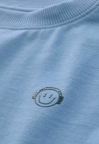 T-shirt en coton bleu clair avec un col rond à côtes, ornée d'un graphique texturé d'un smiley noir portant des écouteurs sur le devant.