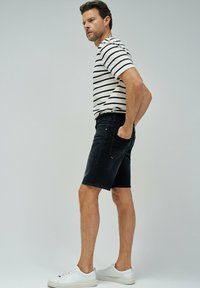 Shorts de mezclilla negra con un corte ajustado y bolsillos laterales, combinados con una camiseta de rayas en blanco y negro. El modelo está de pie con las manos en los bolsillos.