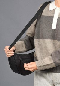Bolso cruzado negro con una textura suave, forma curva y un bolsillo con cremallera. La correa es ajustable; la modelo lleva un suéter a rayas.