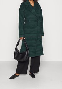 Cappotto a quadri verdi con vita con cintura, abbinato a una borsa nera a tracolla. Scarpe nere basse completano l'outfit.