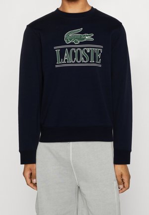 Person iført en sort sweatshirt med en grøn krokodillelogo og teksten "LACOSTE", kombineret med lysegrå bukser.