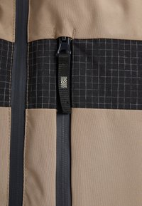 Detailný záber na zips béžovo-čiernej kárované bundy s čiernym ťaháčikom, ktorý má biely vlnitý štvorcový logo.