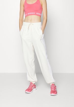 Kobieta w różowym sportowym staniku Adidas, białych spodniach dresowych Adidas z paskami i różowych trampkach Adidas stoi ze włożonymi do kieszeni dłońmi.