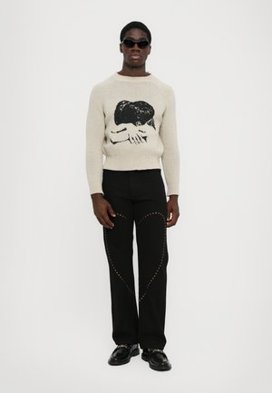 Pull en maille crème avec un design graphique noir, associé à un pantalon noir avec un motif de cœurs cloutés dorés et des chaussures noires brillantes.