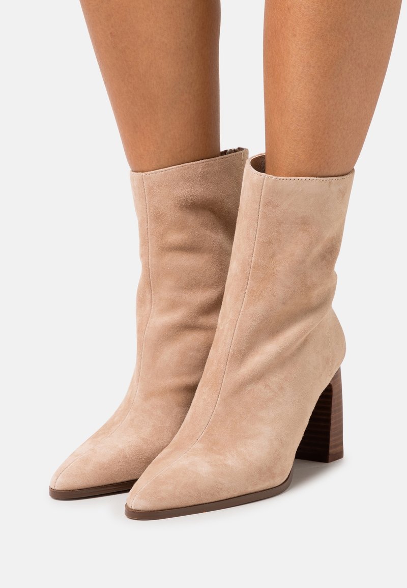 Bottes cheville en daim beige avec bouts pointus et talons blocs empilés portées sur jambes nues sur fond blanc.