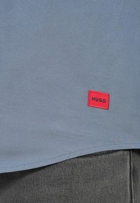 Φωτεινό γαλάζιο πουκάμισο με στρογγυλεμένο τελείωμα και κόκκινη ορθογώνια ετικέτα με ραμμένο το "HUGO" με μαύρα γράμματα. Το ύφασμα φαίνεται λείο με ελάχιστη υφή.