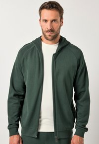 Grüne Zip-Hoodie mit strukturiertem Finish, Raglanärmeln und einem Frontreißverschluss. Wird über einem hellen Hemd getragen und verfügt über eine Kapuze mit Zugband.