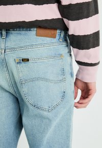 Jeans de denim azul claro con un bolsillo trasero que presenta una etiqueta negra de "Lee". Textura suave, corte clásico y un parche de cuero marrón en la cinturilla.