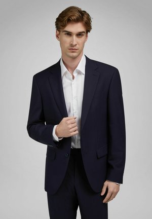 Młody mężczyzna poprawiający granatową marynarkę na białej koszuli, stojący na tle jednolitego szarego tła, o poważnym wyrazie twarzy.