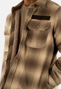 Camisa de franela a cuadros marrón con forro de forro polar, que cuenta con dos bolsillos frontales, botones a presión y un parche texturizado de "VON DUTCH" en el pecho.
