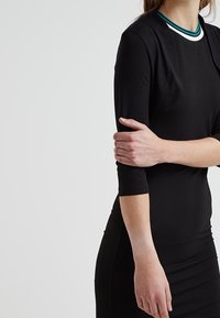 Svart klänning med trekvartslånga ärmar, rund halsringning med grön och vit kant, figurnära design, slät tygtextur.