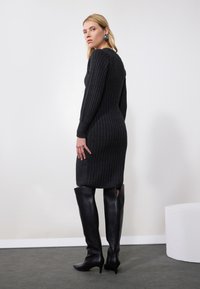 Ribbed mustat neulemekko pitkillä hihoilla, tiukka siluetti, hieman polven ylle ulottuva pituus, yhdistettynä korkeisiin mustiin nahkasaappaisiin.