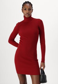 Sarkans ribots turtlenecks kleita ar garām piedurknēm, pieguļošs siluets un vidēja augstuma garums līdz vidus augšstilbam. Apvienota ar mazu melnu rokassomu.