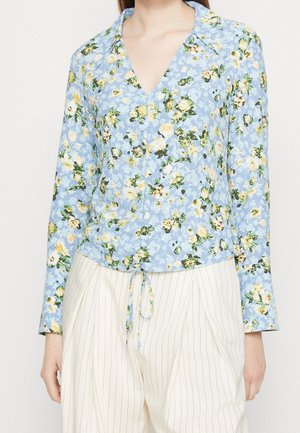 Vrouw draagt een blauwe bloemenprint blouse met lange mouwen, kraag en strik aan de voorkant, gecombineerd met witte, hoogtallige broek met fijne strepen.