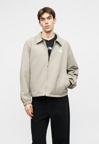 Νεαρός άντρας με κοντά καστανά μαλλιά, φορώντας ανοιχτό μπεζ μπουφάν με φερμουάρ πάνω από μαύρο πουκάμισο και μαύρο παντελόνι, στέκεται μπροστά σε απλό φόντο.