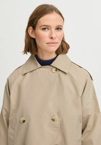 Manteau trench beige à double boutonnage avec de gros boutons, un large col et des manches raglan. Tissu lisse et léger avec une coupe décontractée.
