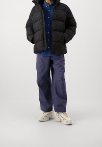 Giacca a vento nera con motivo paisley, camicia blu con bottoni, pantaloni cargo viola e calzature beige con ritagli.