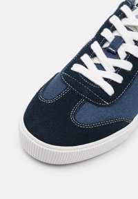 Zapatillas de deporte azul marino que presentan una mezcla de ante y tela, cordones blancos, costuras de contraste y una suela de goma texturizada.