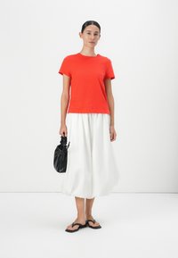 Tricou roșu cu mânecă scurtă asociat cu o fustă albă vaporoasă, o geantă texturată neagră și sandale negre, având un design simplu, minimalist.