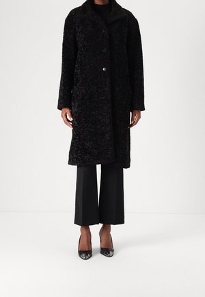 Personne portant un long manteau noir texturé, un pantalon large noir et des talons hauts noirs à bouts pointus, debout devant un fond blanc uni.