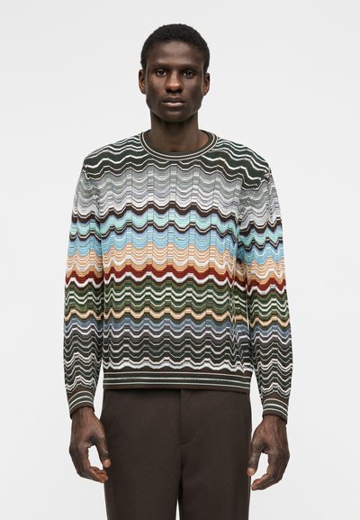 Άντρας που φοράει πουλόβερ με πολύχρωμο ζιγκ ζαγκ μοτίβο σε αποχρώσεις του πράσινου, μπλε, καφέ και μπεζ, συνδυασμένο με σκούρο καφέ παντελόνι.