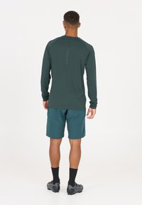 Mörkgrön långärmad t-shirt för träning med rund halsringning och raglanärmar; kombinerad med turkosa shorts och svarta strumpor.