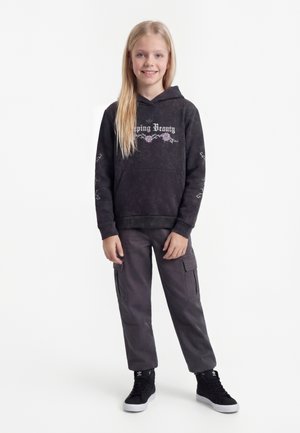 Fille aux longs cheveux blonds debout, souriante, vêtue d'un sweat à capuche noir "La Belle au bois dormant", d'un pantalon cargo foncé et de baskets montantes noires.