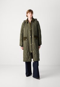 Gequiltede olijfgroene jas met capuchon, voorzien van gouden hardware, corduroy accenten en twee voorkruiden. Gekocht over flared denim broeken.