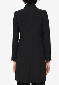 Frau mit lockigem Haar, die einen eng anliegenden schwarzen knielangen Mantel mit langen Ärmeln trägt, von hinten vor einem weißen Hintergrund gesehen.