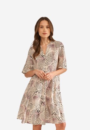 Woman with long brown hair wearing a knee-length beige dress with black and pink abstract animal print, standing with hands gently clasped.