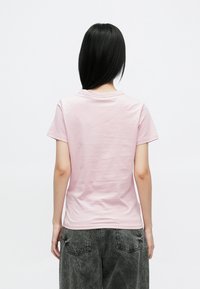 Kortærmet, lyserød T-shirt vist bagfra. Materialet virker glat. Rund hals med diskret syning. Kombineret med grå bukser med høj talje.