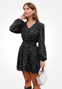 Abito nero con motivo floreale bianco, vita arricciata, maniche a palloncino e scollatura a V. Abbinato a una borsa bianca e una cintura in evidenza.