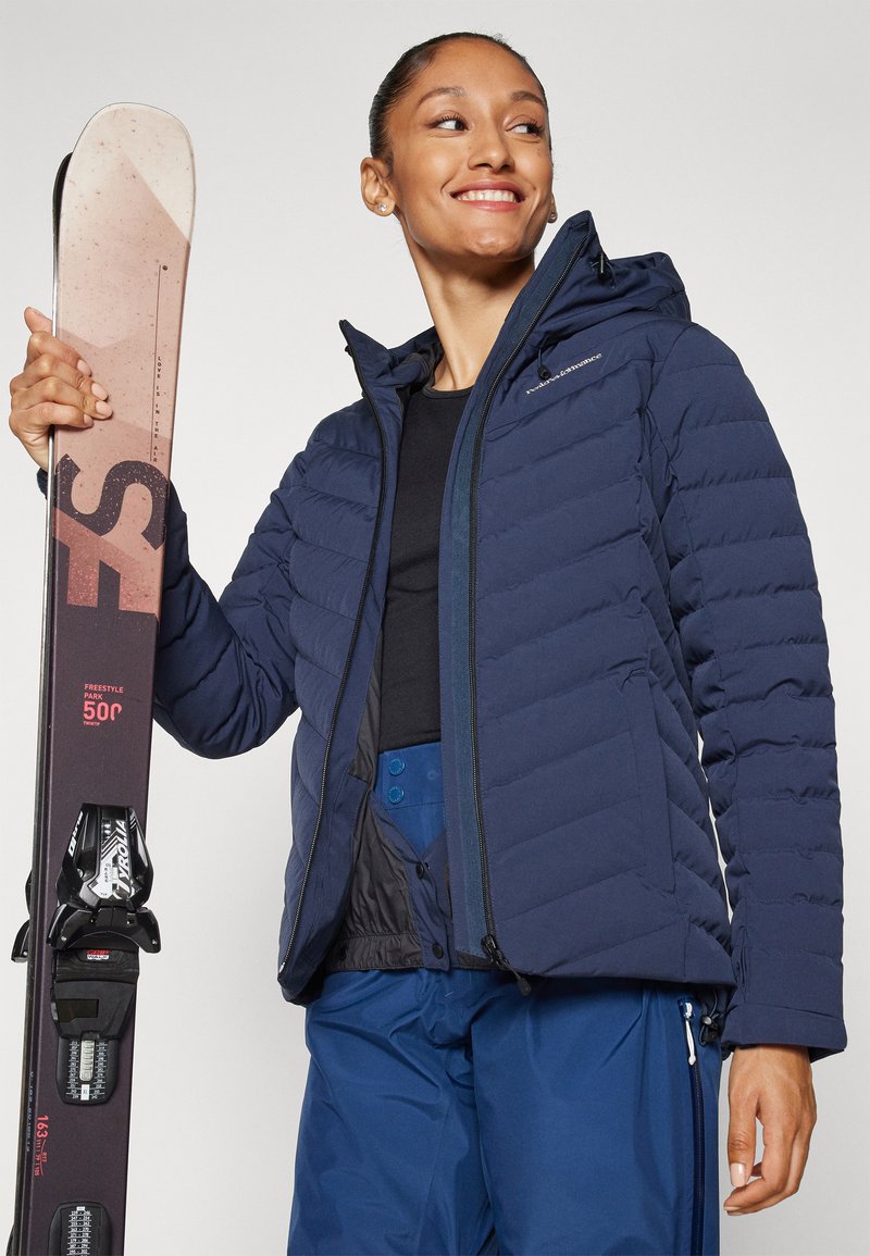 Marineblaue Puffjacke mit horizontaler Steppung und einer anliegenden Kapuze. Das Modell hält einen pinkfarbenen Ski mit schwarzen Details. Sie trägt außerdem blaue Skihosen.