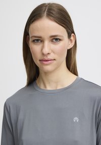 Langarm, lichtgrijs sportshirt gemaakt van gladde stof met een klein wit logo op de linkerborst en een ronde hals.